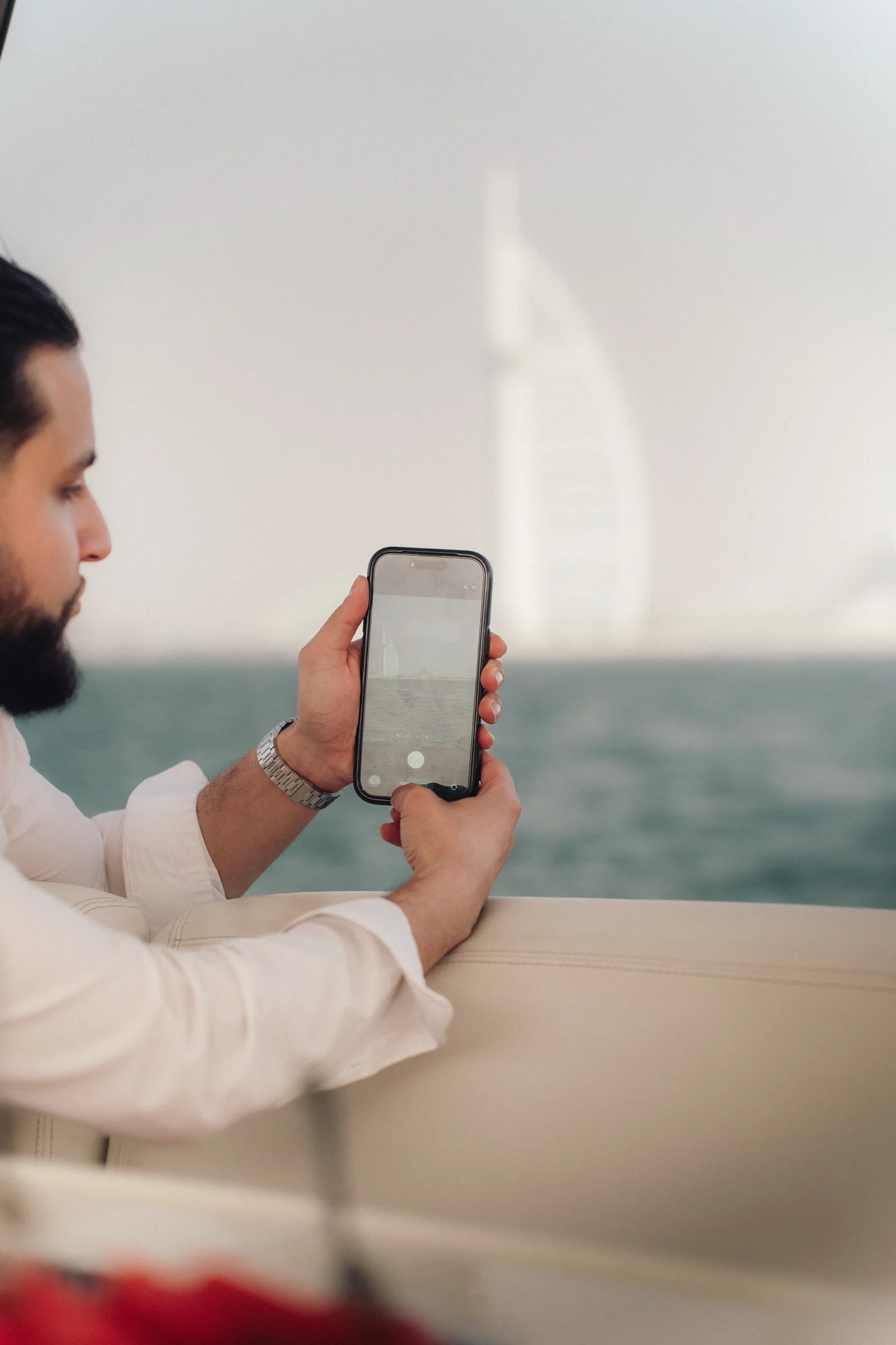 Zack Lalouti holding up his phone to photograph the Dubai waterfront from a boat, with the Burj Al Arab in the distance.