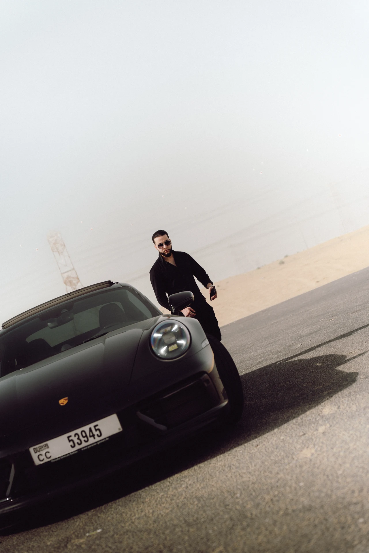 Zack Lalouti wearing sunglasses beside the front of a black Porsche on a desert road in Dubai.