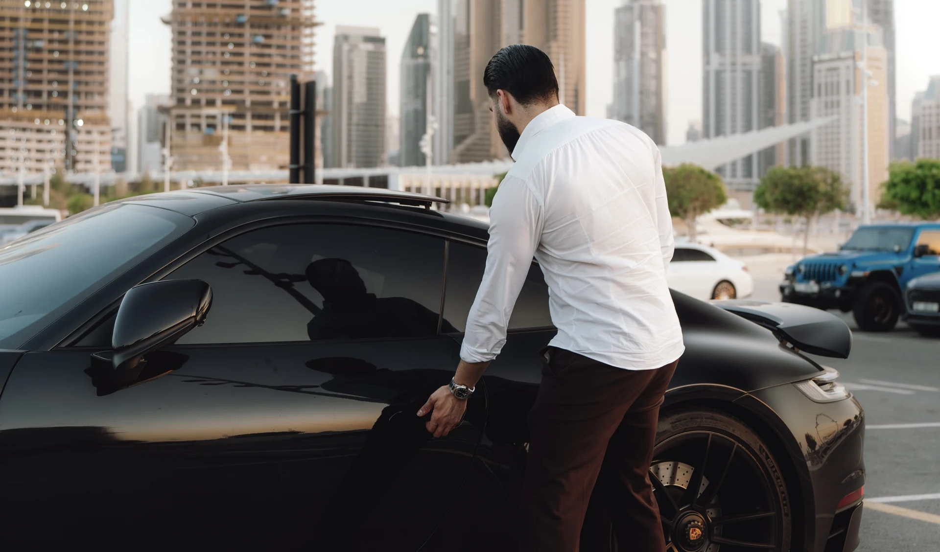 Zack Lalouti wearing a white shirt beside the door of a black Porsche, with high-rise buildings in the background.